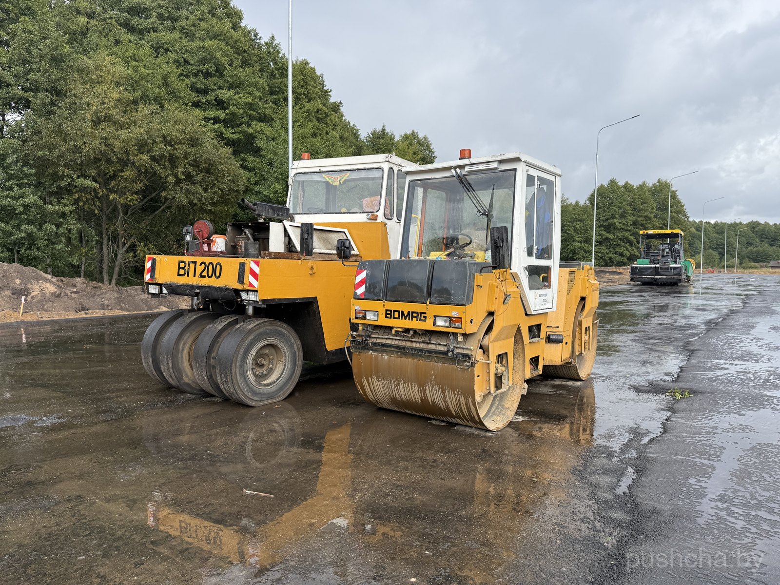 Два дорожных катка BOMAG крупным планом (вибровалец и пневмоколесный «ВП 200»); мокрый асфальт, фонари, лес позади.