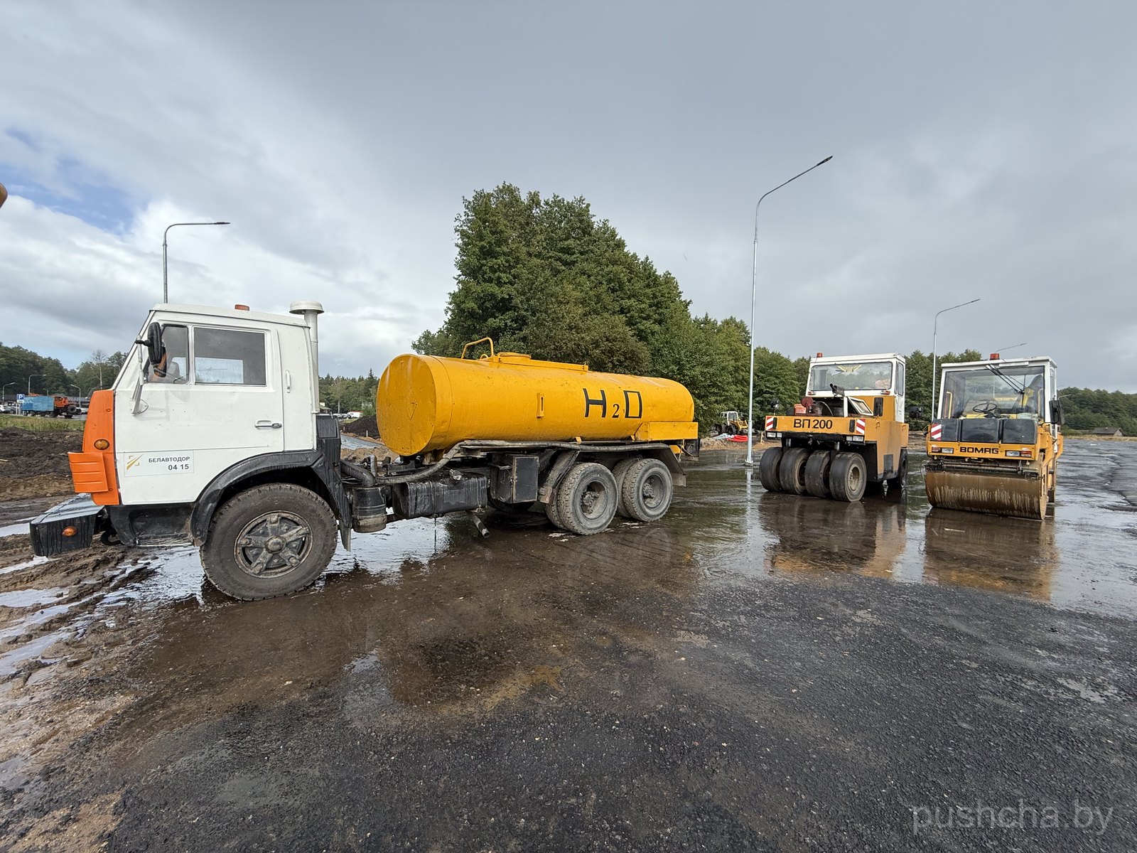 Белый водовоз на грунтовом участке слева; справа два катка (ВП 200 и BOMAG) на зеркальном асфальте; граница грязь/асфальт.